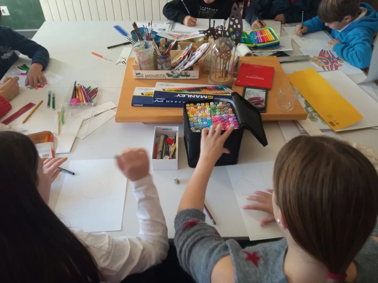 Nens i nenes dibuixant al voltant de la taula de l’estudi, amb retoladors i llapis